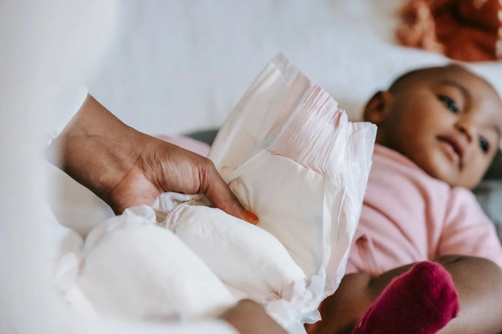 Mão de adulto segura uma fralda aberta enquanto um bebê de blusa rosa, deitado ao lado, olha para ela na hora da troca