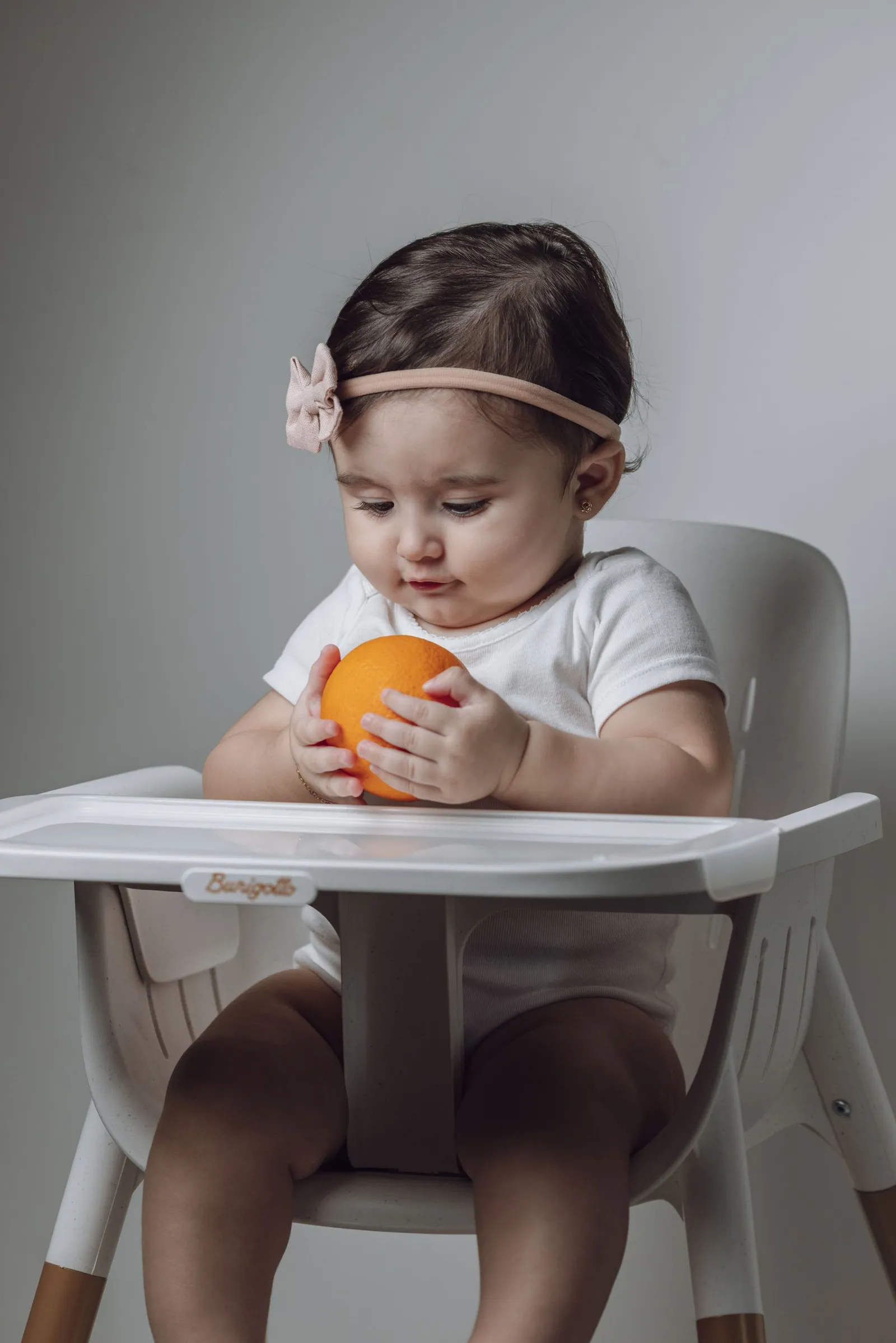 Bebê em cadeira alta segurando uma laranja, com faixa de cabelo, em retrato de estúdio com fundo cinza