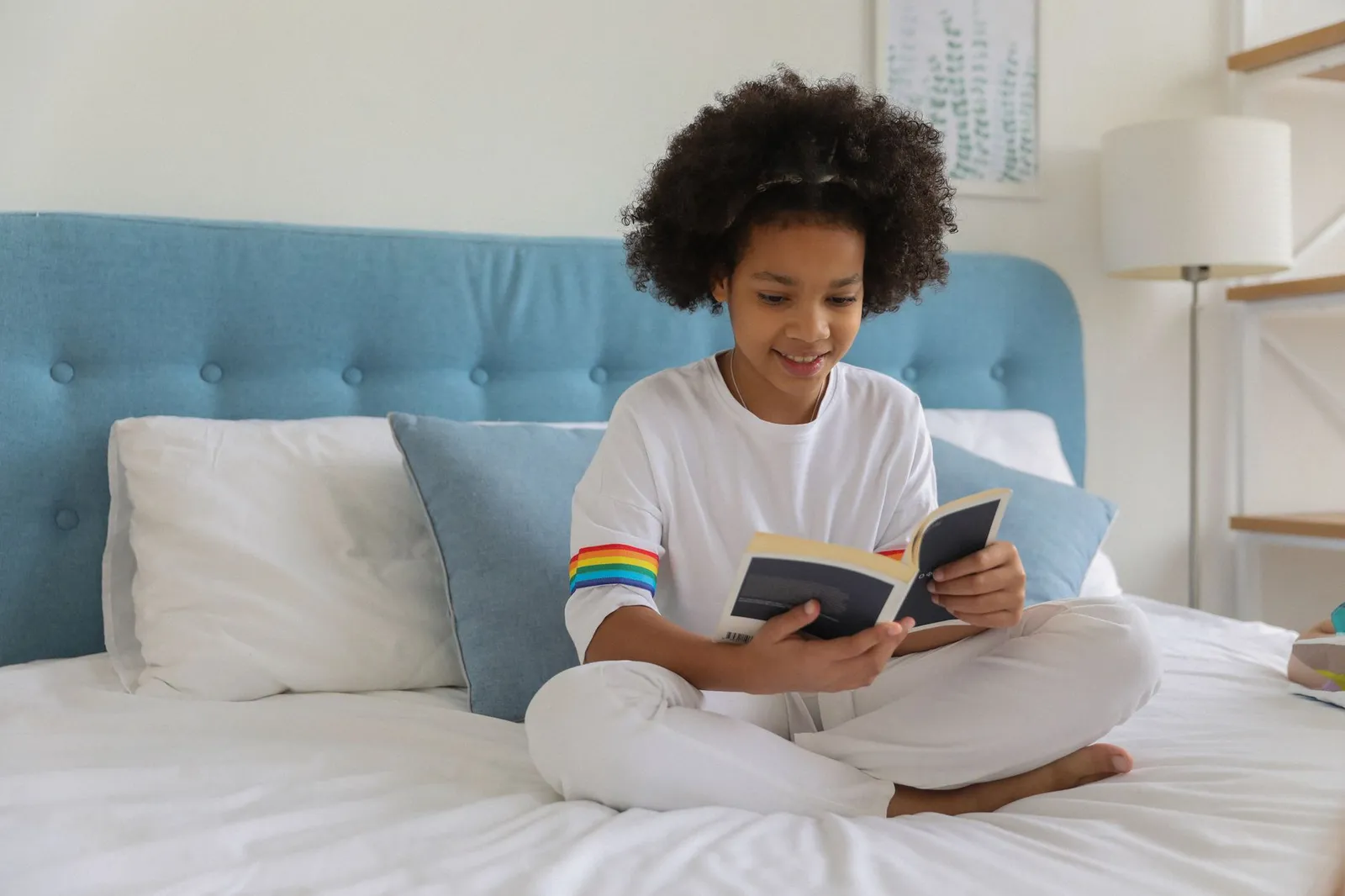 Menina sorrindo enquanto lê um livro de capa dura sentada na cama em quarto claro