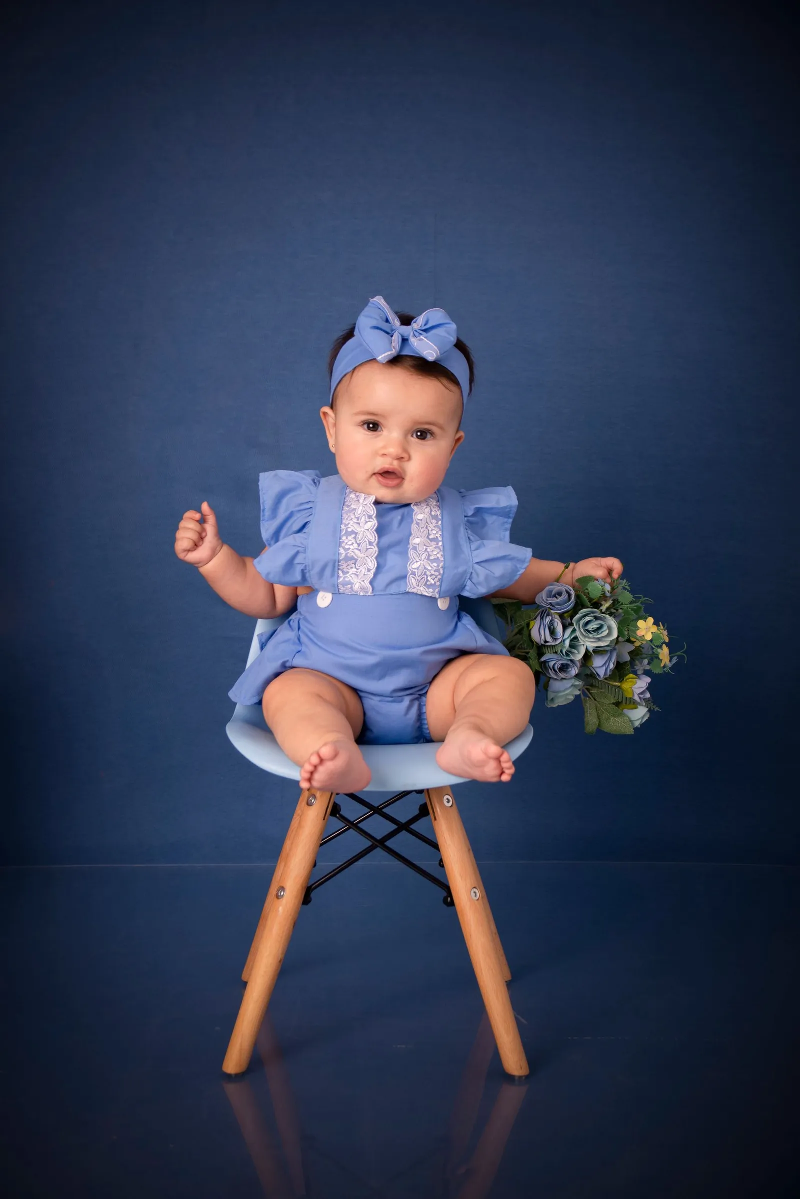 Bebê menina em traje azul sentada em cadeira decorada com flores em estúdio fotográfico
