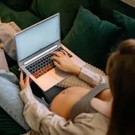 Gestante sentada em sofá verde usando um notebook, foto vista de cima, na Calculadora de Gravidez da Mamii