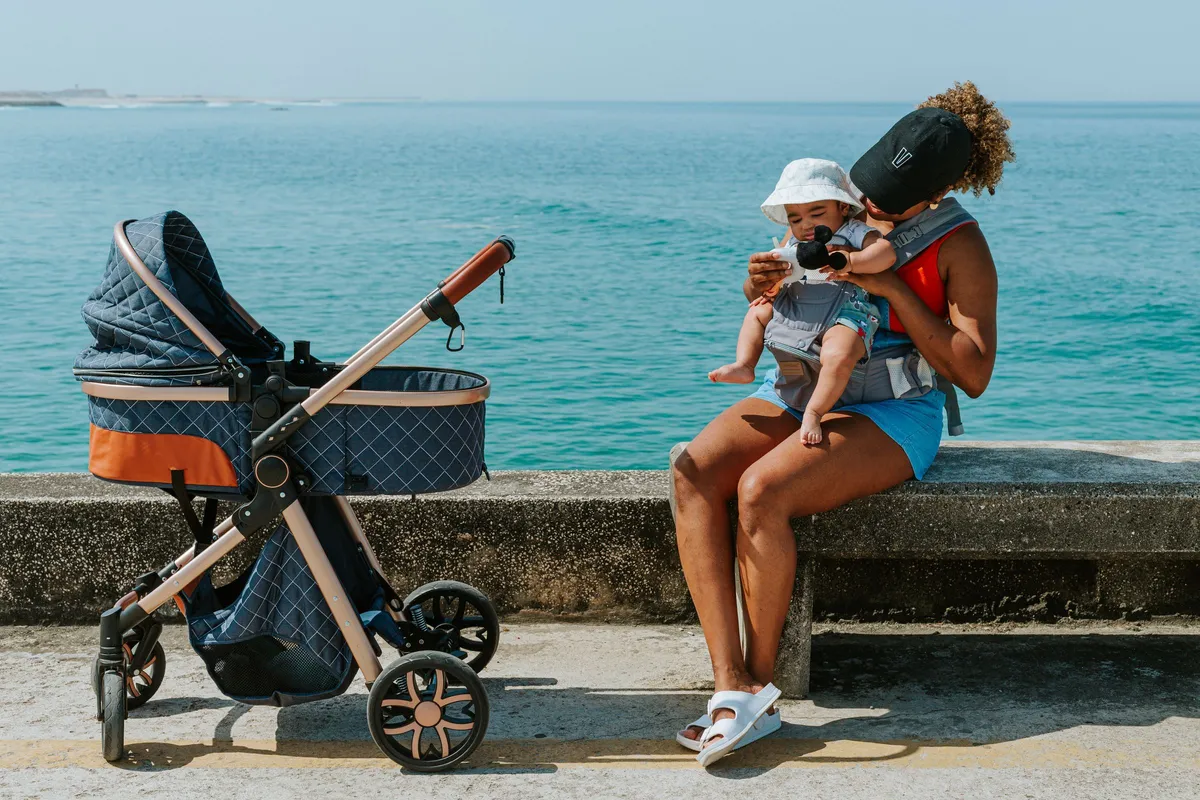 Mãe com o bebê no canguru sentada em uma mureta à beira do mar, ao lado de um carrinho azul, na categoria Passeio e Viagem