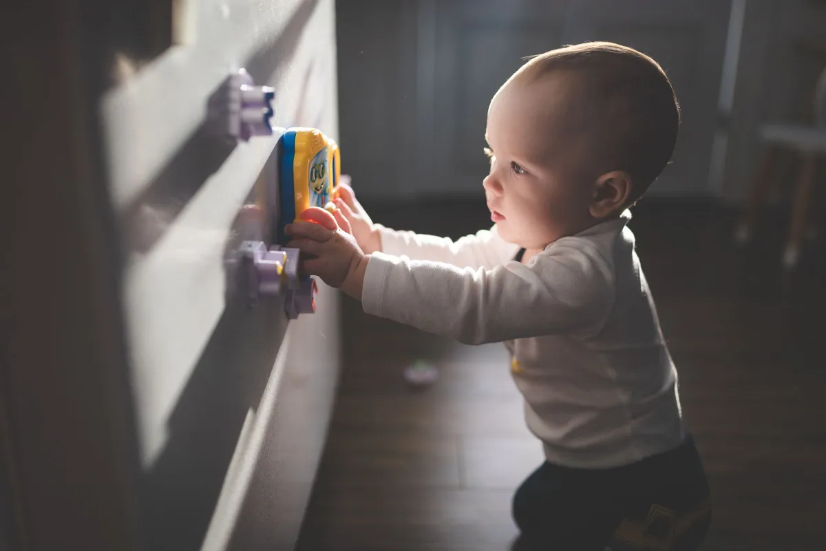 Bebê em pé, de perfil, apoiado na parede e segurando um brinquedo amarelo e azul, na categoria Segurança do Bebê
